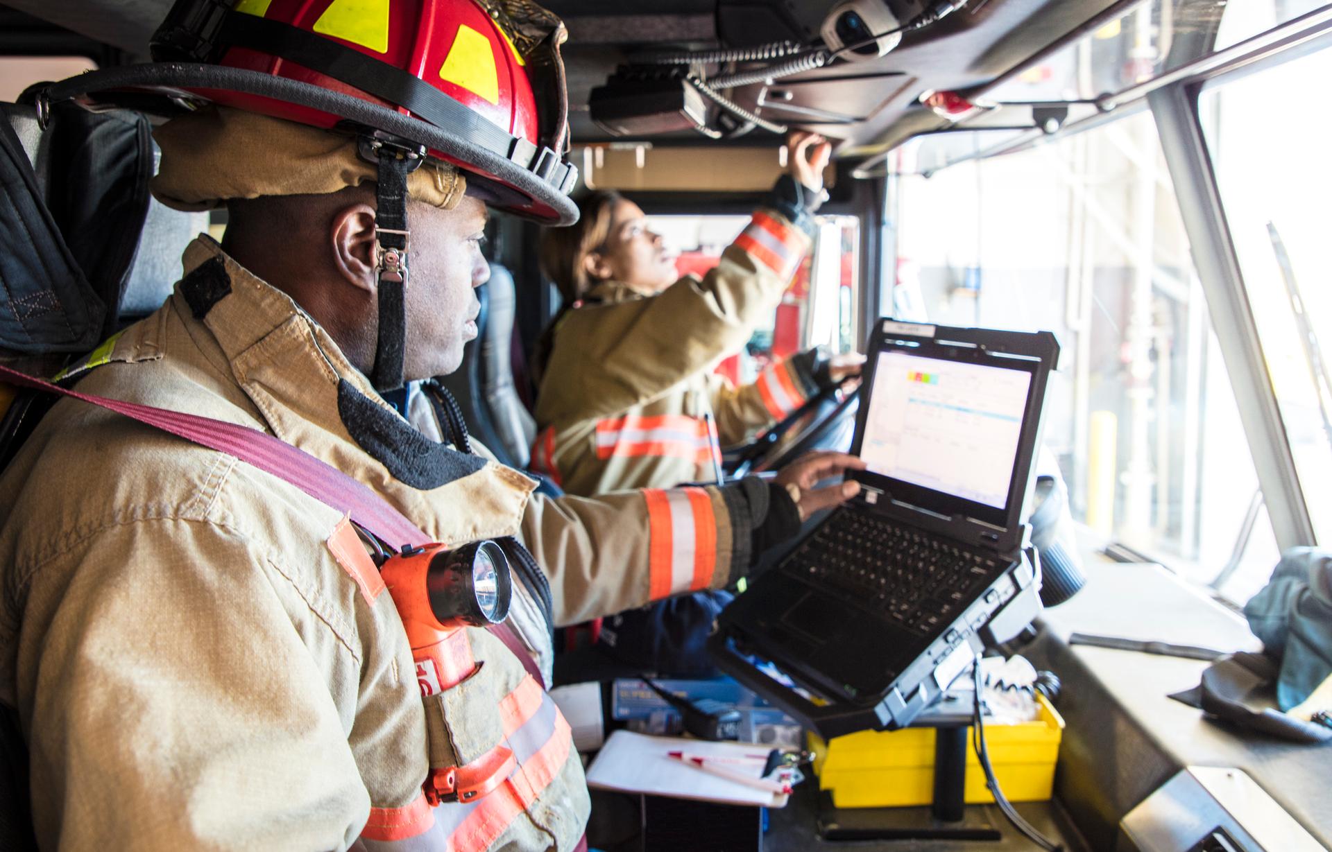 Two firefighters sitting in the firetruck looking at a laptop.